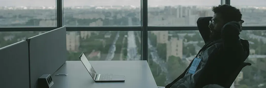 Man sitter avslappnat vid skrivbord med dator framför sig och blickar ut över staden – symboliserar trygghet genom skydd av personuppgifter som personnummer.