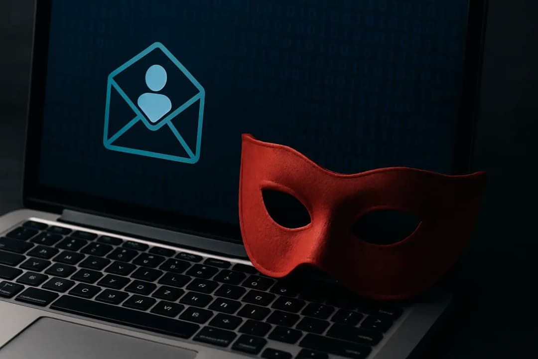 Ultra-realistisk bild av en laptop med ett rött maskeradmask bredvid tangentbordet och ett e-postikon med användarsymbol på skärmen – symboliserar digital identitetsstöld och spoofing i cybersäkerhetssammanhang.